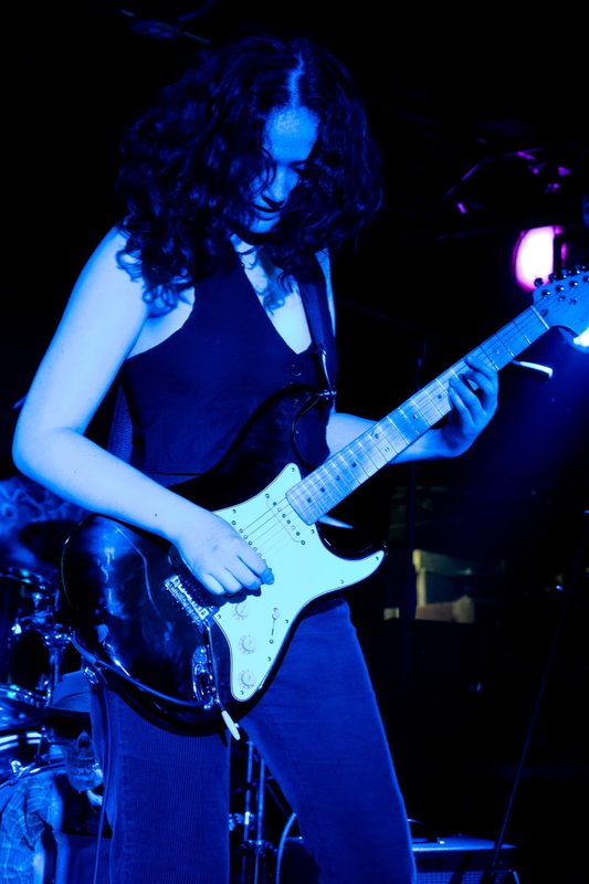 Kylee under blue stage light, looking down at a black Stratocaster