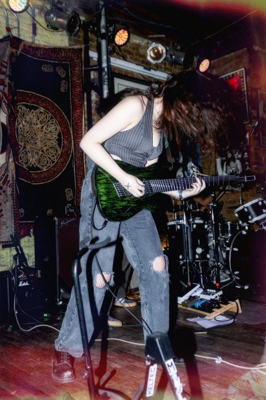 Kylee headbanging mid-riff on a green seven-string guitar in a brick-walled club