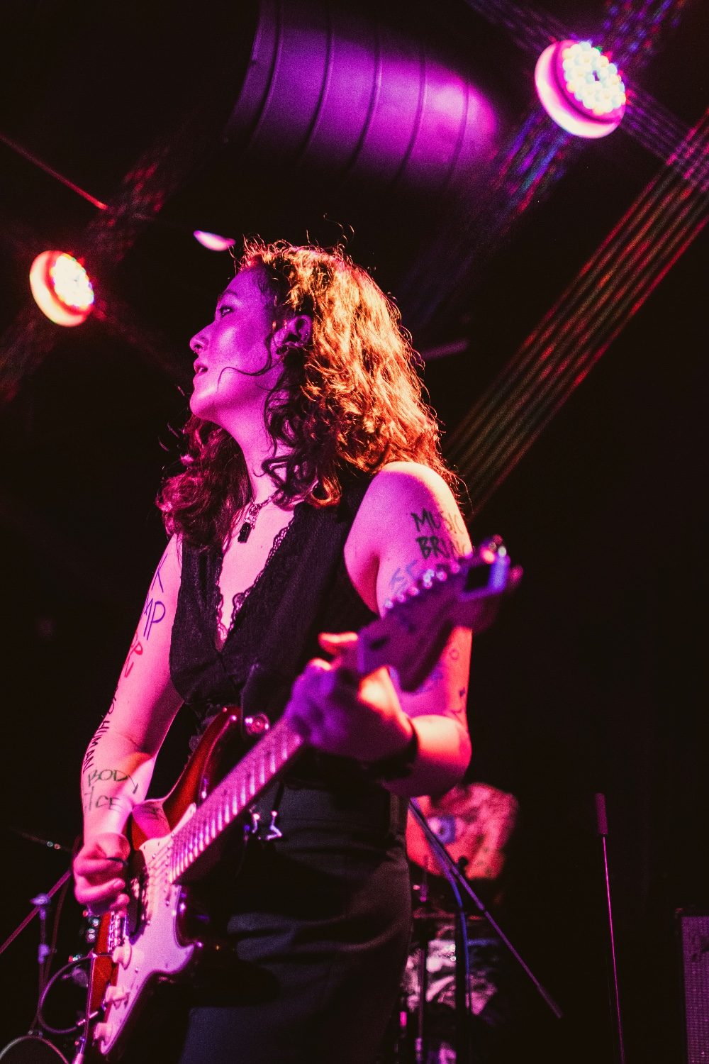 Kylee on stage in pink and red light, looking out past the lighting rig while playing a red Stratocaster