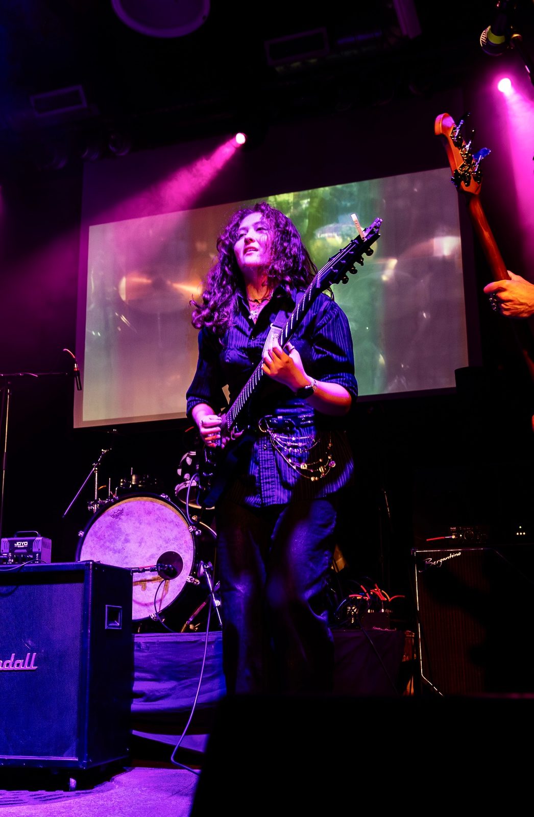 Kylee on a purple-lit stage with a seven-string guitar, a projection screen glowing behind