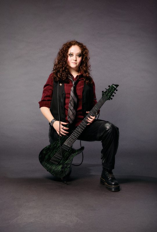 RED STATE promo shot: Kylee kneeling with a green seven-string guitar, in a red shirt, tie and black vest
