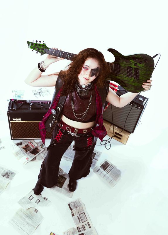 RED STATE promo shot: Kylee in star face paint holding a green seven-string guitar behind their head, amps and newspapers around them