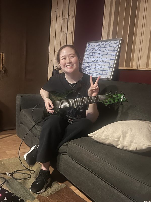 Kylee in a recording studio on a couch with a green seven-string guitar, smiling and throwing a peace sign
