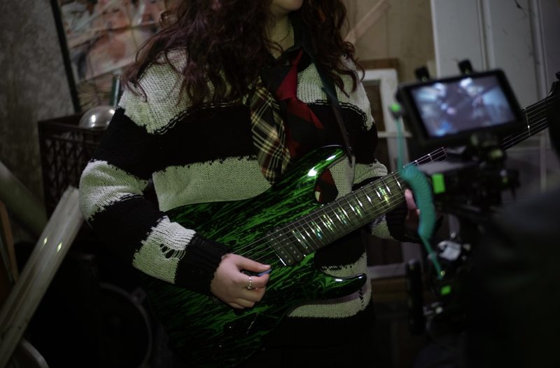 Close-up of Kylee playing a green seven-string guitar on a video shoot, camera rig in the background
