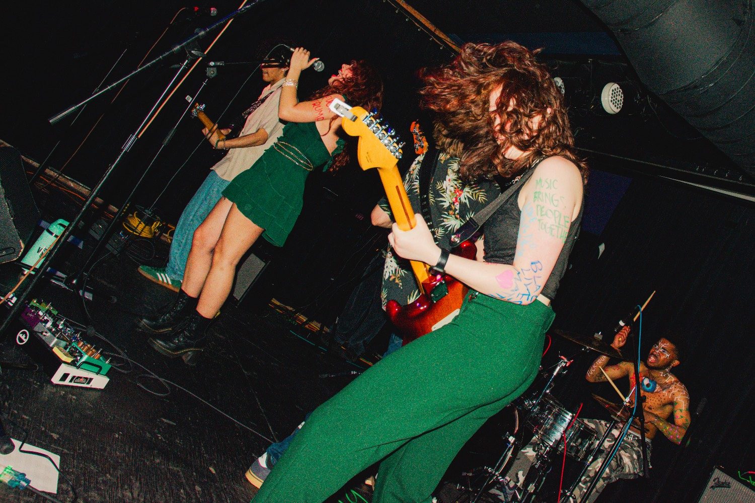 Versus Vitality live: Kylee headbanging with a red guitar in front, singer in a green dress, painted drummer behind